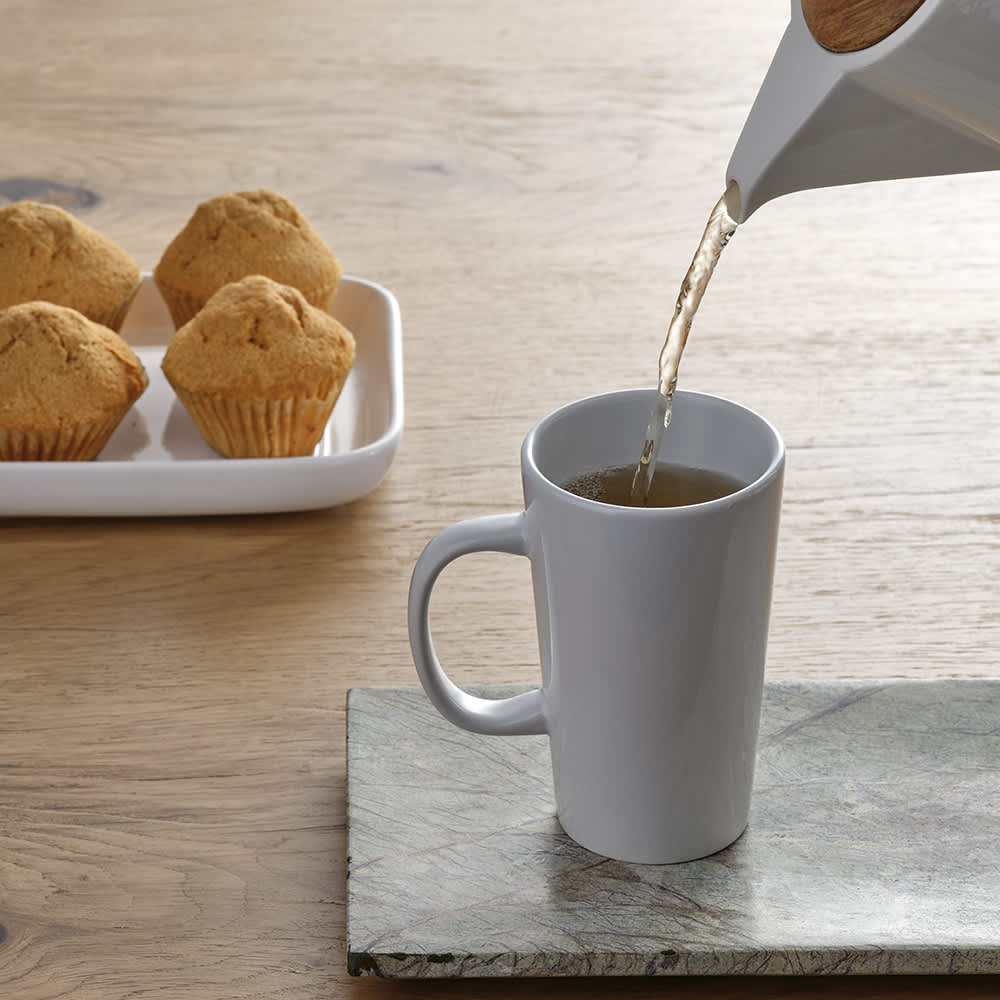 A white ceramic mug being filled with tea, alongside a tray of muffins on a wooden surface.