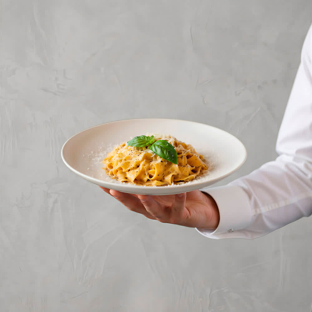 A ceramic dinner plate with a matt finish, featuring pasta topped with basil leaves. The plate is held by a person in a white shirt.