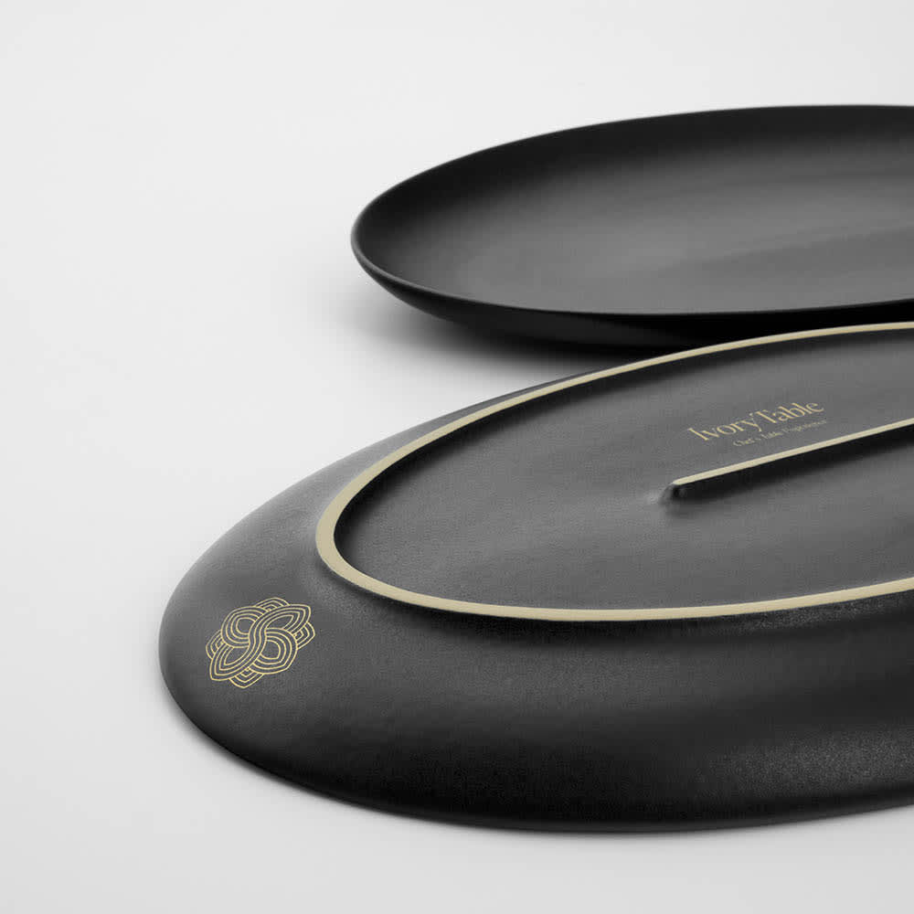 A matte black ceramic platter with a decorative edge, featuring a logo and a smooth, elegant design.