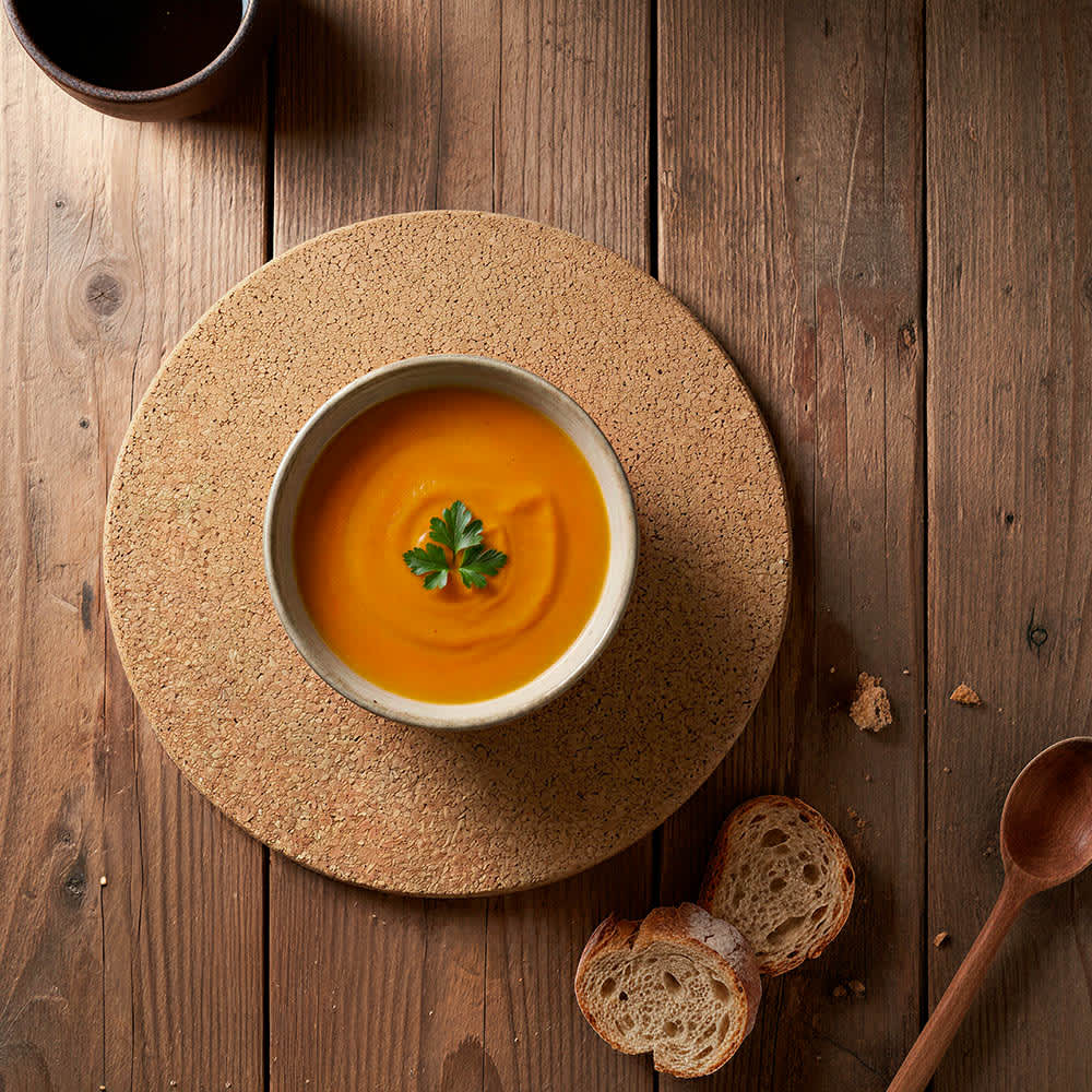 A round cork placemat in natural tones supports a bowl of orange soup, garnished with a green herb, on a wooden table.