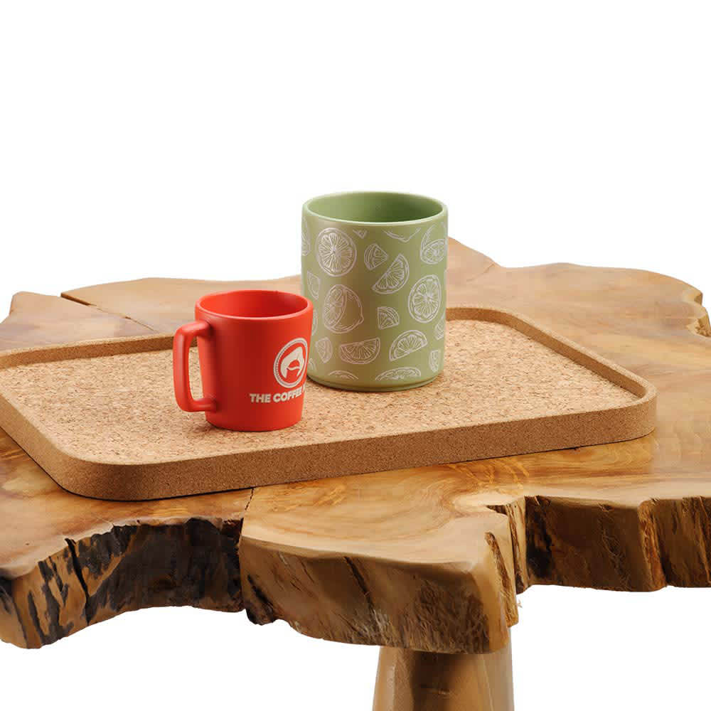 A rectangular cork tray in natural tones holds a red mug and a green mug, placed on a rustic wooden surface.