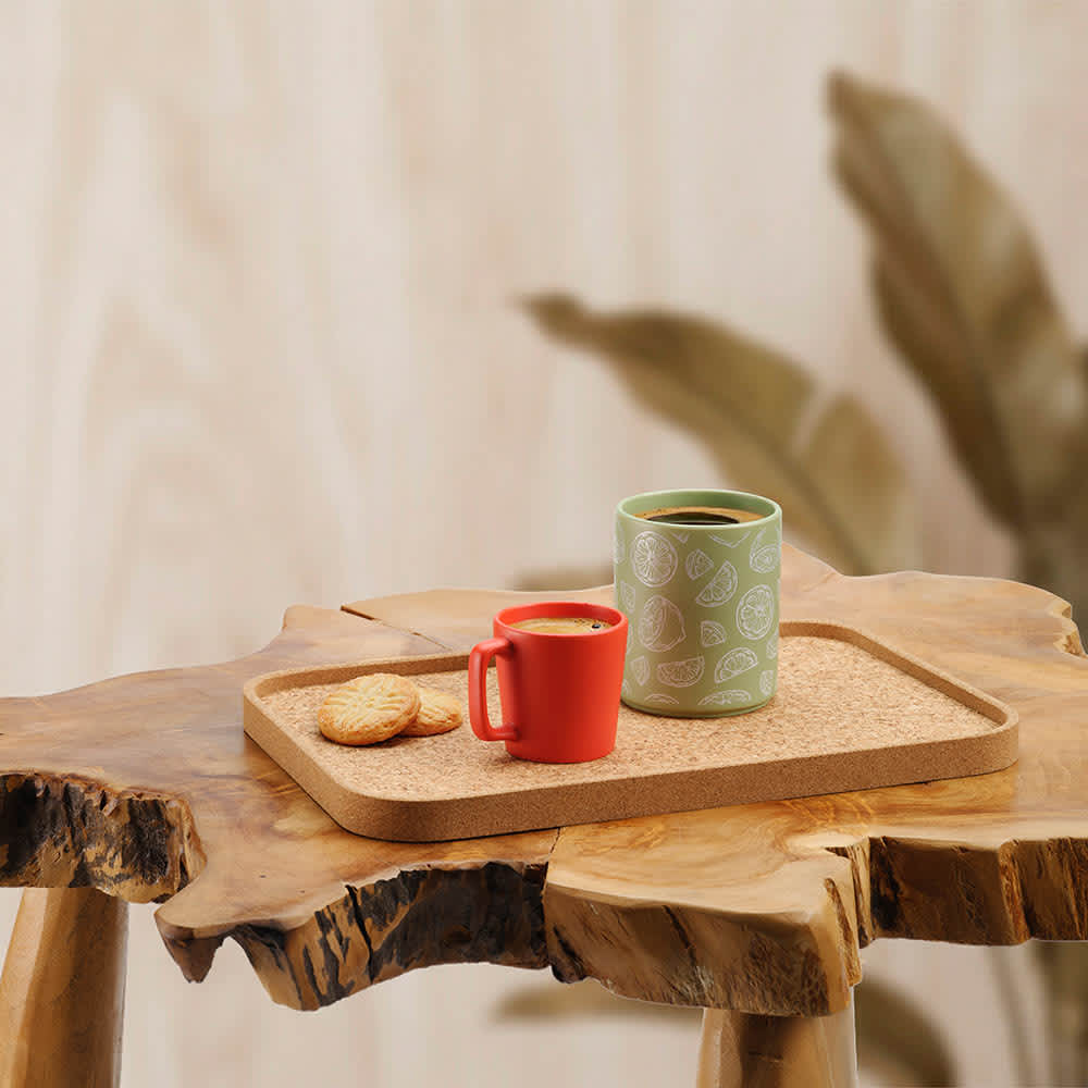 A rectangular cork tray in a natural finish, featuring a red cup, a green mug, and some biscuits on a wooden table.
