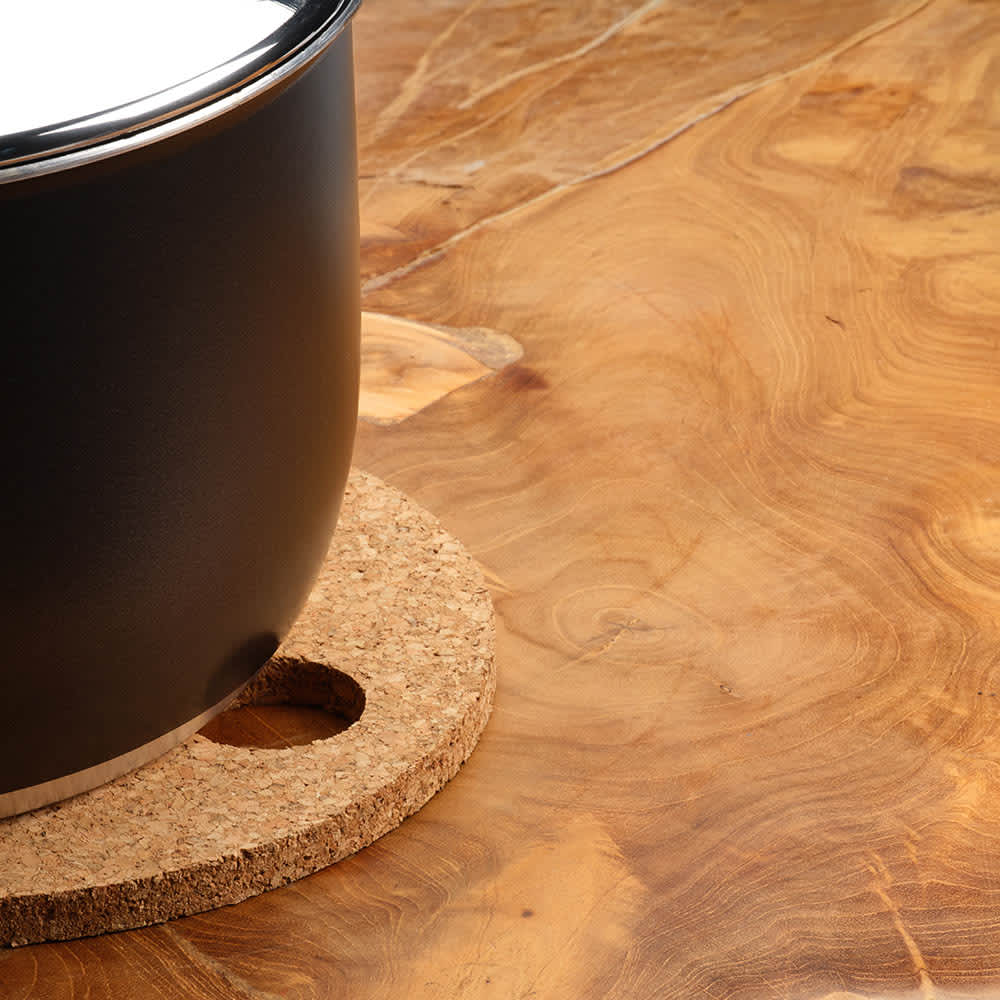A round cork trivet in a natural finish, placed under a black pot on a wooden surface.