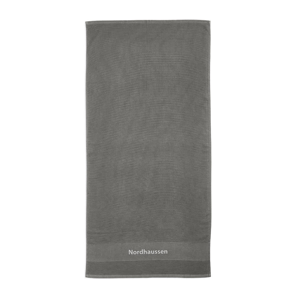 A grey towel made of cotton, featuring a subtle texture and a logo at the bottom. The towel is thick with a 500 gsm weight.