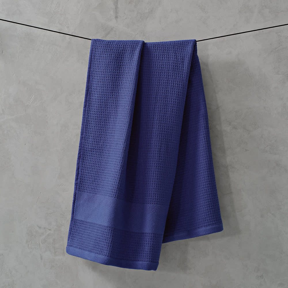 A purple cotton towel with a textured pattern, hanging against a grey background. It features a logo.