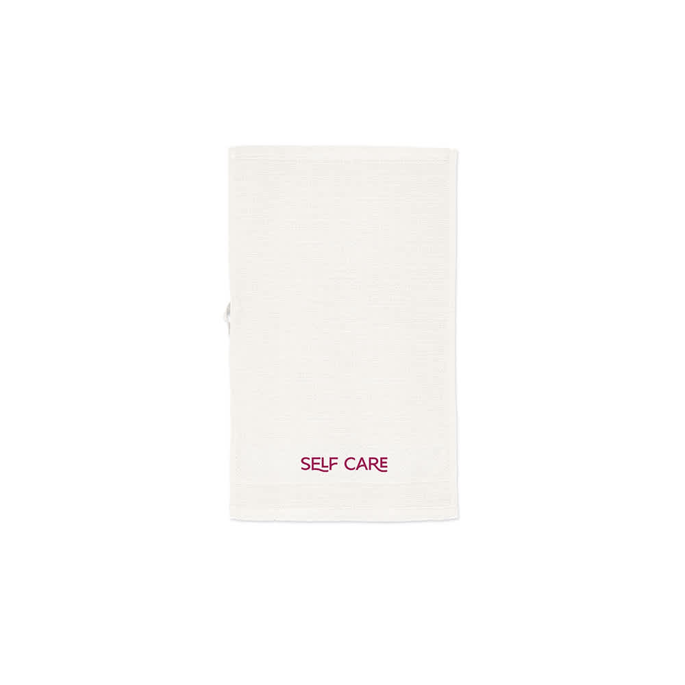 A cream-coloured towel made of cotton with a branding logo, featuring the words "SELF CARE" in burgundy.