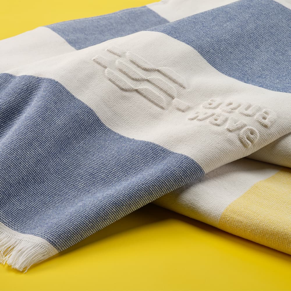 A cotton towel featuring a striped pattern in blue, white, and yellow, with a textured logo.