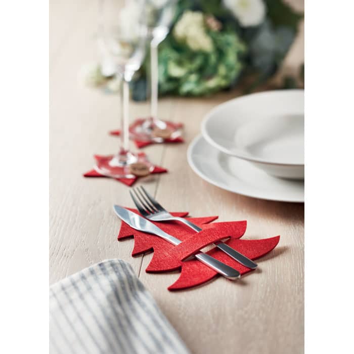 A red felt cutlery holder shaped like a Christmas tree, placed on a wooden table with plates and glasses.