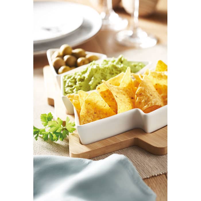A snack platter featuring corn chips, guacamole, and olives in white bowls on a wooden board, with a table setting in the background.