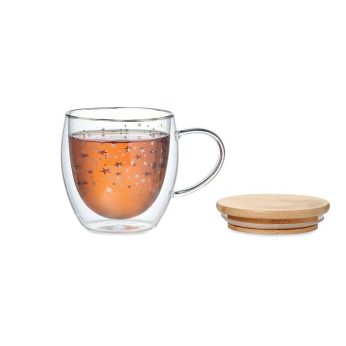A glass mug featuring a star pattern, alongside a round wooden coaster. The mug holds a warm brown beverage.
