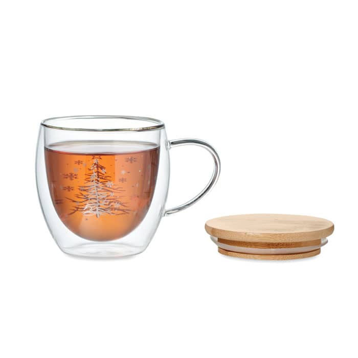 A clear glass cup with a Christmas tree design, filled with brown tea, accompanied by a bamboo coaster.