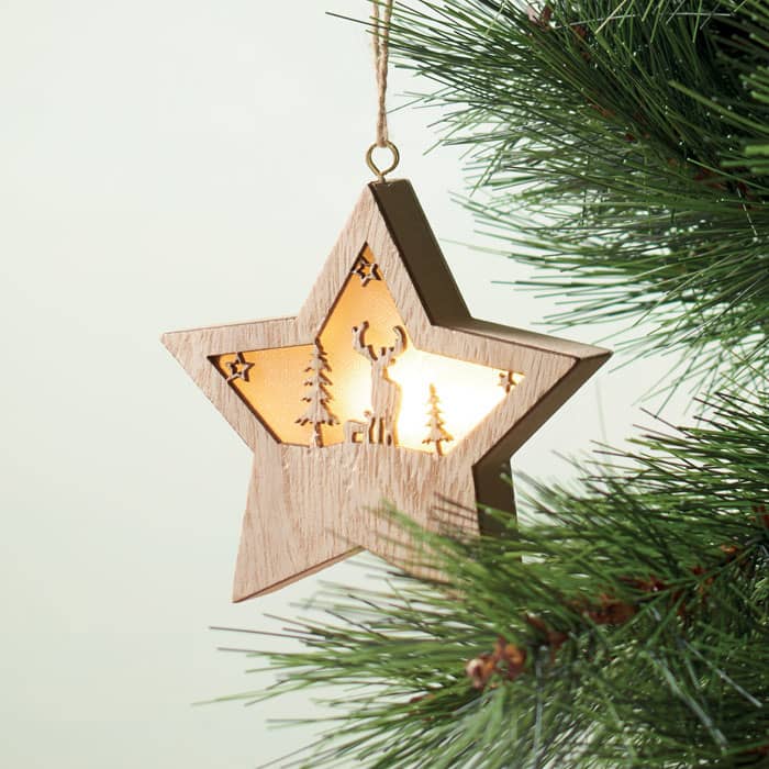A wooden star ornament with a light, featuring cut-out tree designs, hanging from a pine branch.