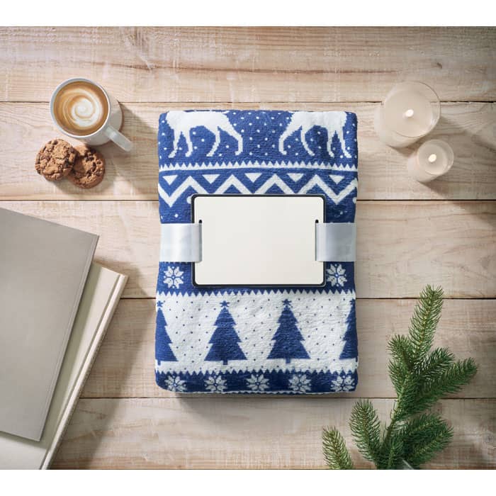 A blue blanket with a winter pattern, placed on a wooden table alongside a cup of coffee, cookies, and candles.