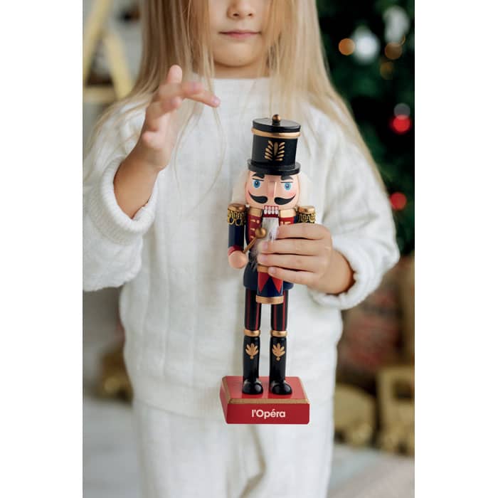 A nutcracker figurine in a military outfit held by a child, featuring red, black, and gold colours against a festive background.