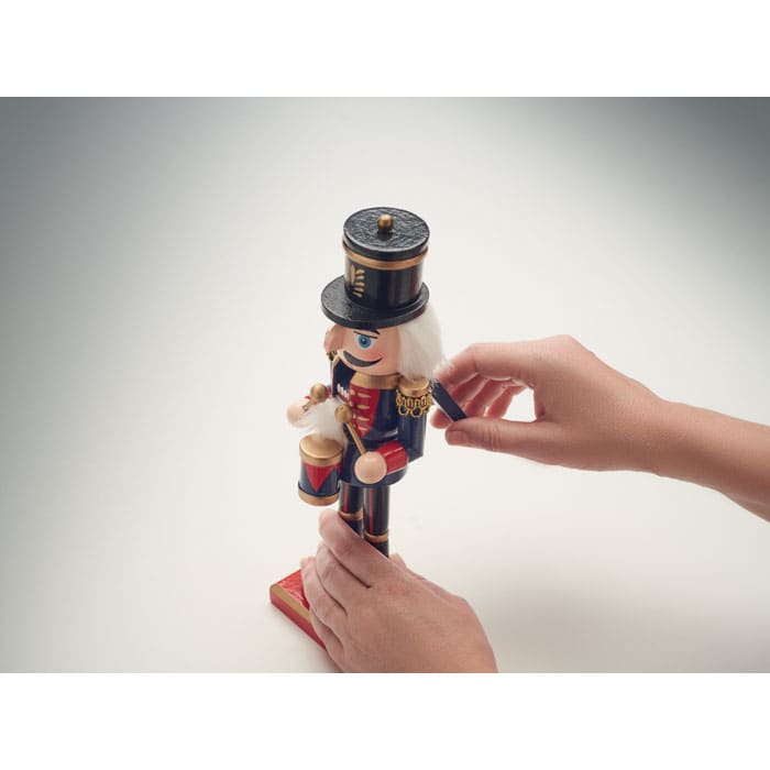 A toy nutcracker figure with a black hat, blue and red uniform, holding a drum, being manipulated by a person’s hand.