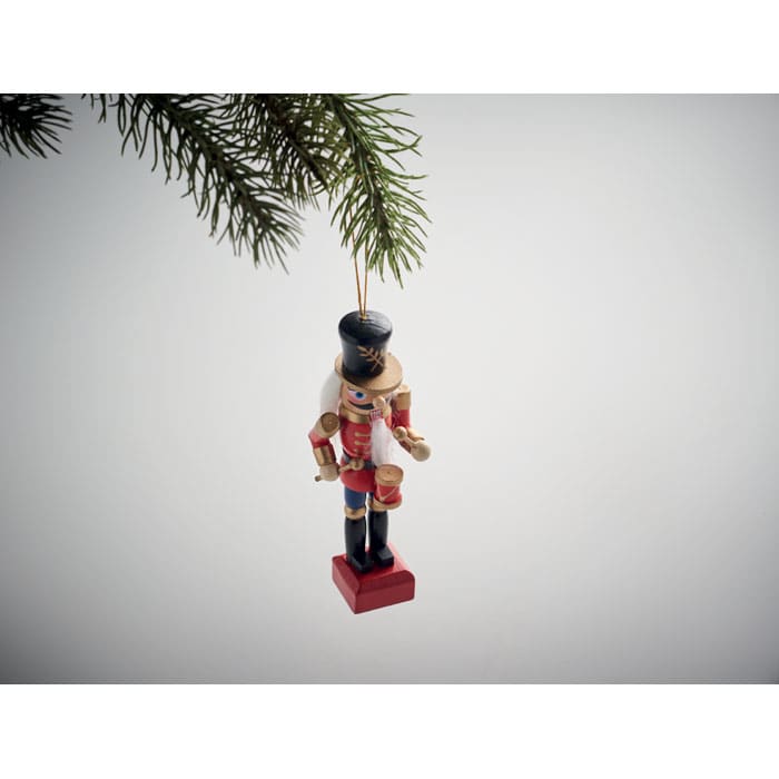 A nutcracker ornament in traditional attire, hanging from a green pine branch against a grey background.
