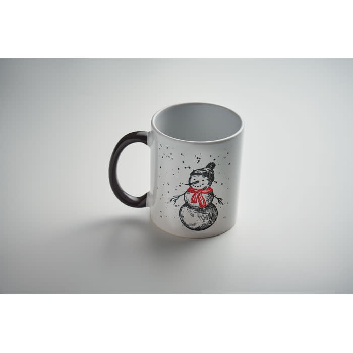A white mug featuring a snowman design, with a black handle and snowy details. It has a logo on the side.