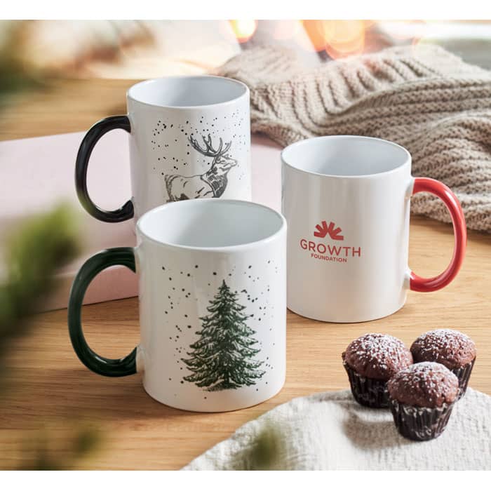 Three mugs in green, white, and red, featuring festive designs, with chocolate treats nearby. One mug has a logo.
