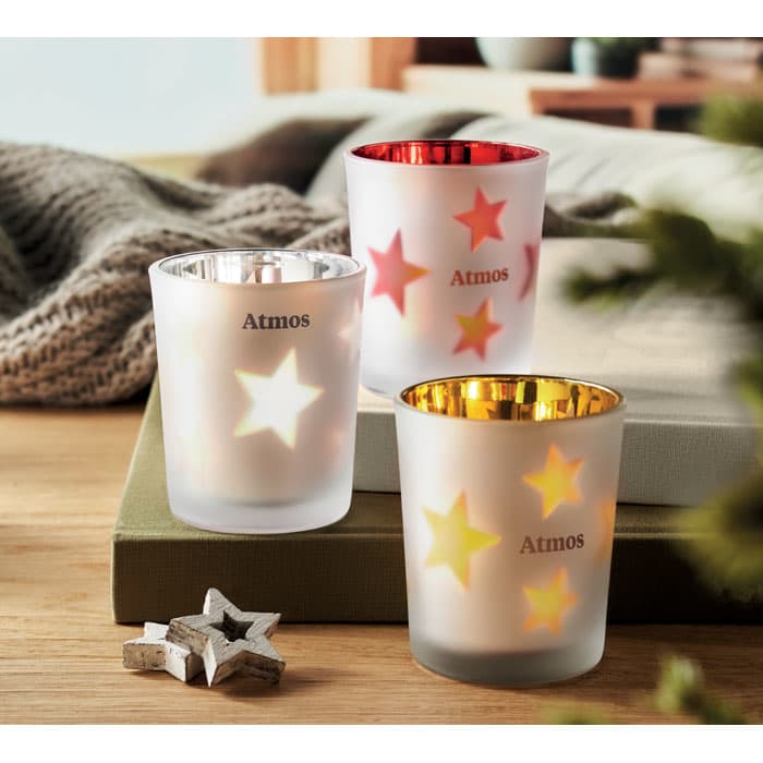 Three candle holders in frosted glass with star cut-outs, featuring red, gold, and clear finishes, placed on a soft surface.