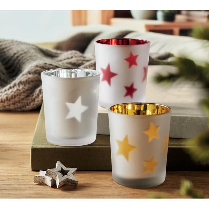 Three decorative glass candle holders in frosted white, red, and gold, featuring star designs on a wooden surface.