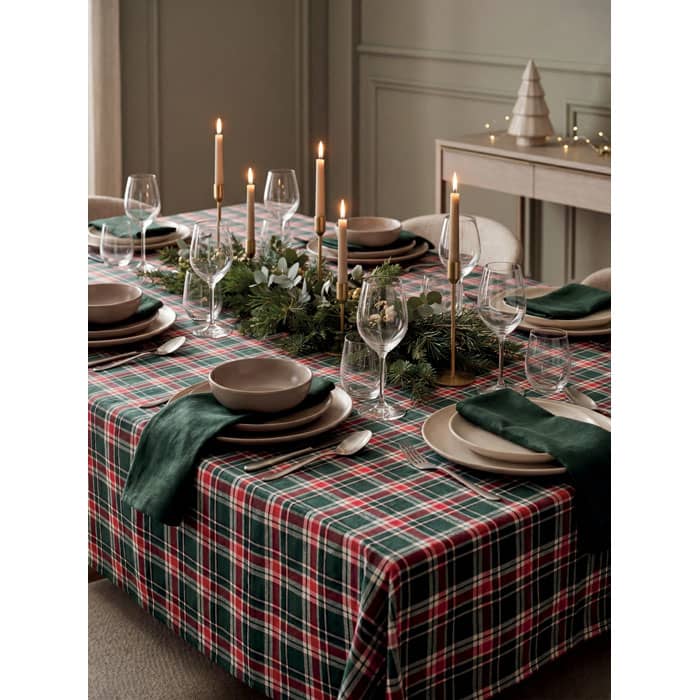 A festive dining table set with a red and green plaid tablecloth, ceramic plates, glassware, and tall candles.
