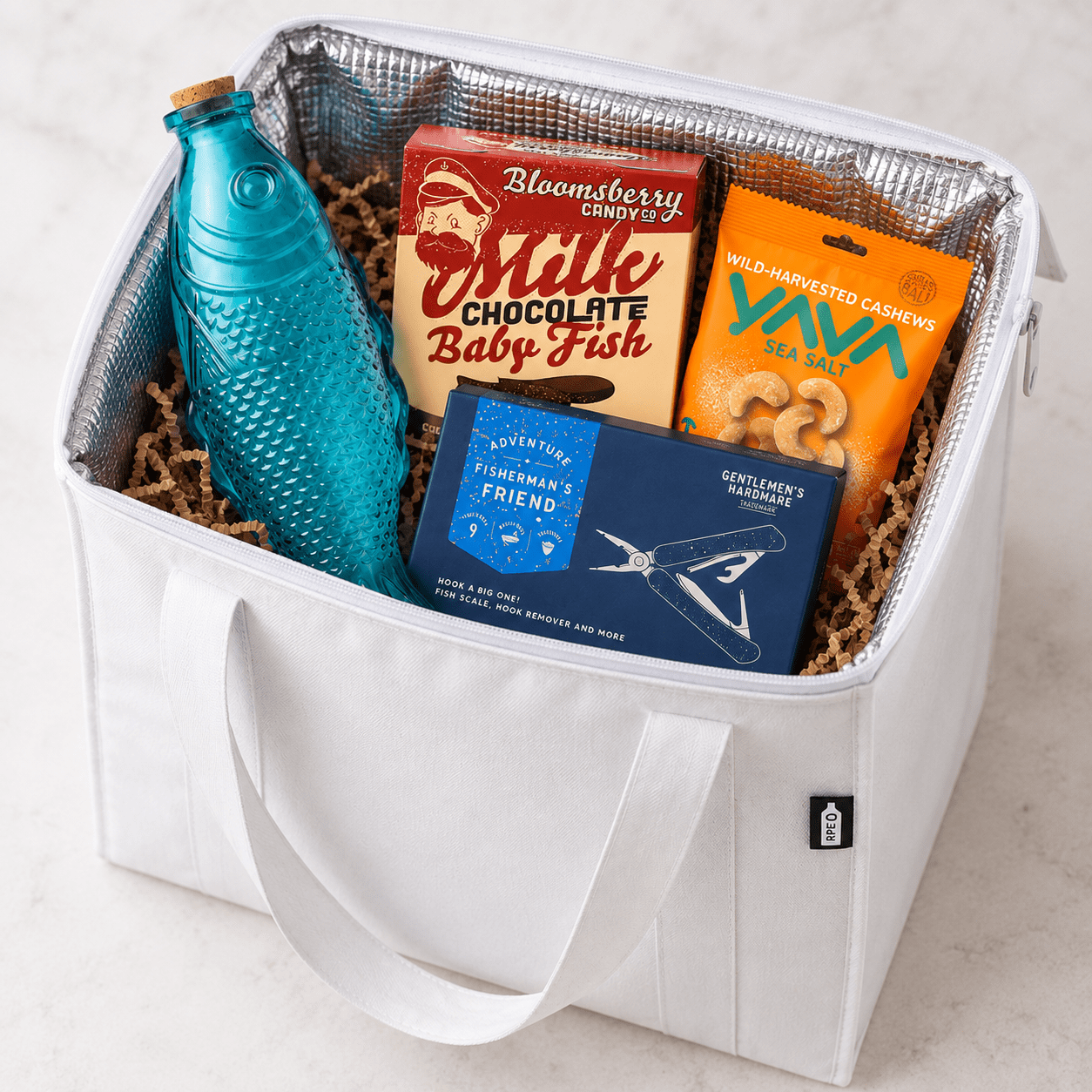 A white cooler bag containing a blue fish-shaped drink bottle, chocolate, sea salt, and fishing-themed items.