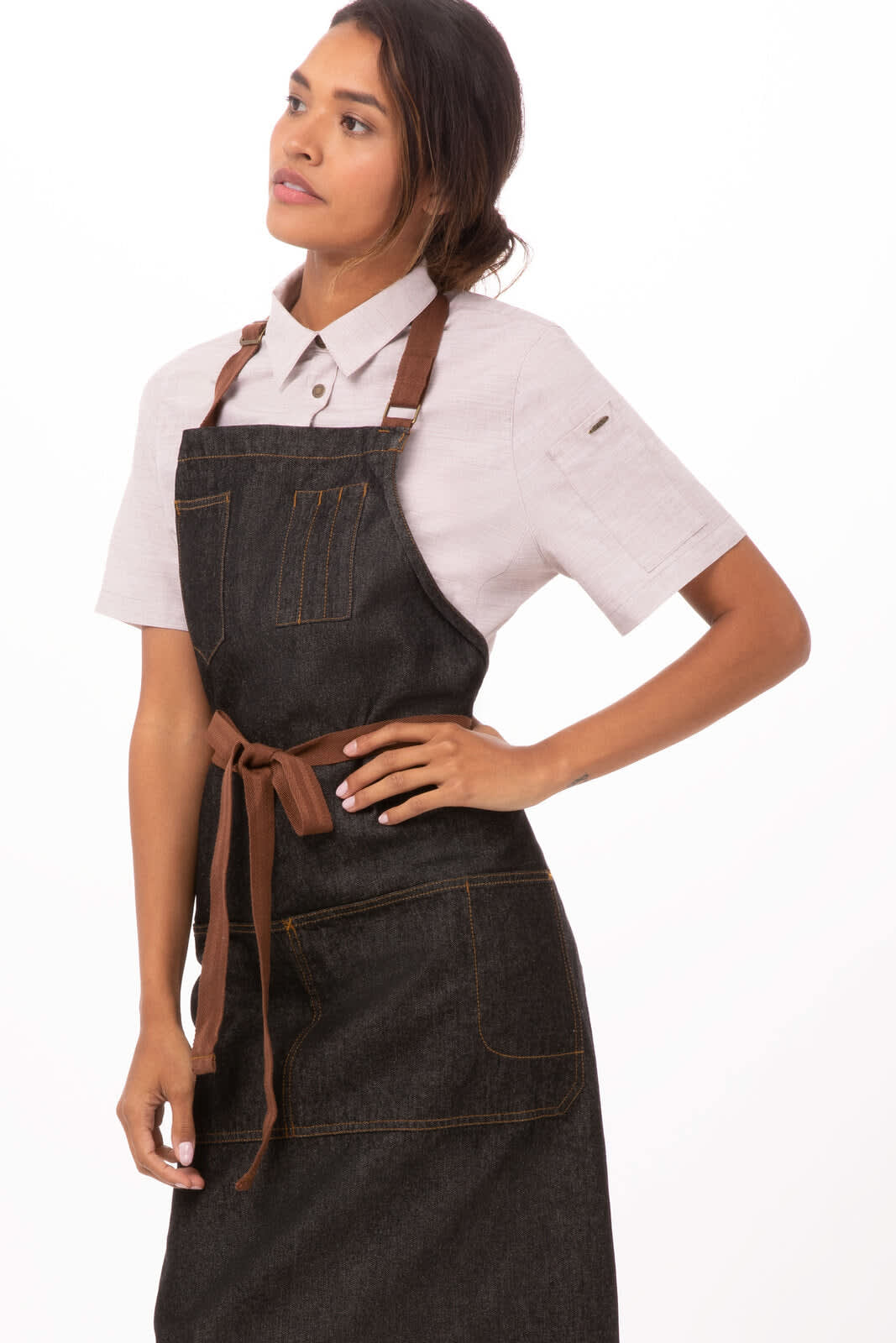 A woman models a dark denim bib apron with brown straps, featuring pockets and an overall stylish design.