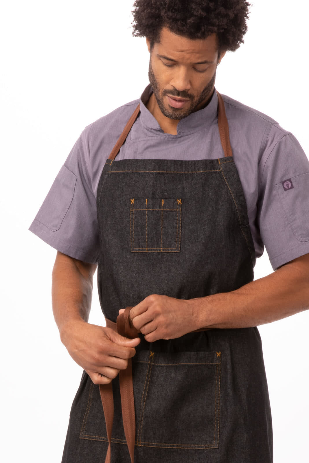 A dark denim bib apron with leather straps, worn by a person adjusting the waist tie. Featuring a logo.
