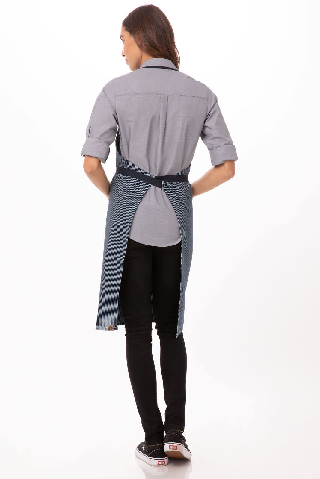 A back view of a denim Portland Bib Apron worn over a grey shirt, featuring a logo. The model has dark pants and shoes.
