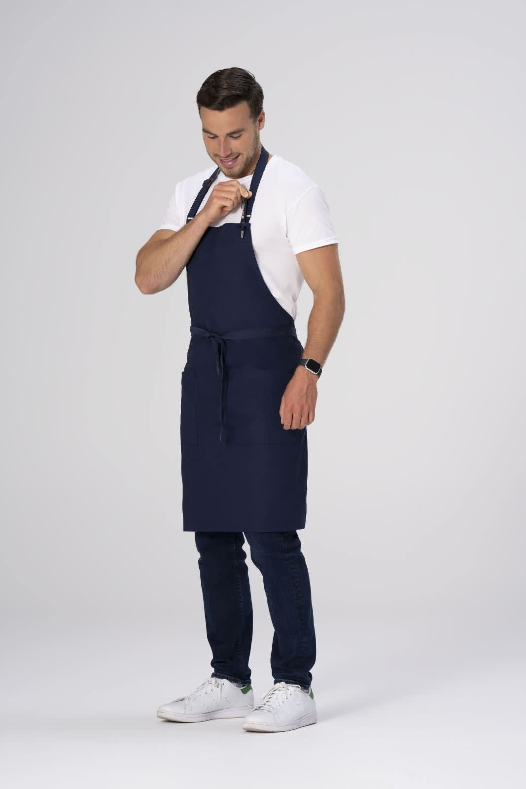 A man in a navy blue apron adjusts it while wearing a white t-shirt and dark jeans. The apron has a branding logo.