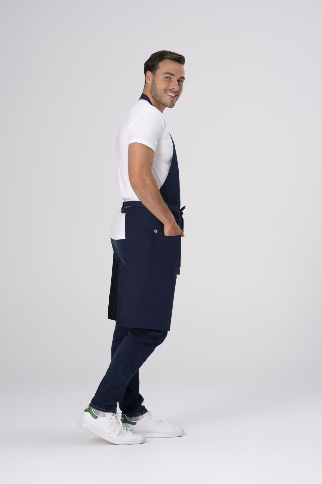 A man wearing a navy Lockharte Bib Apron over a white t-shirt and jeans, standing with a smile.