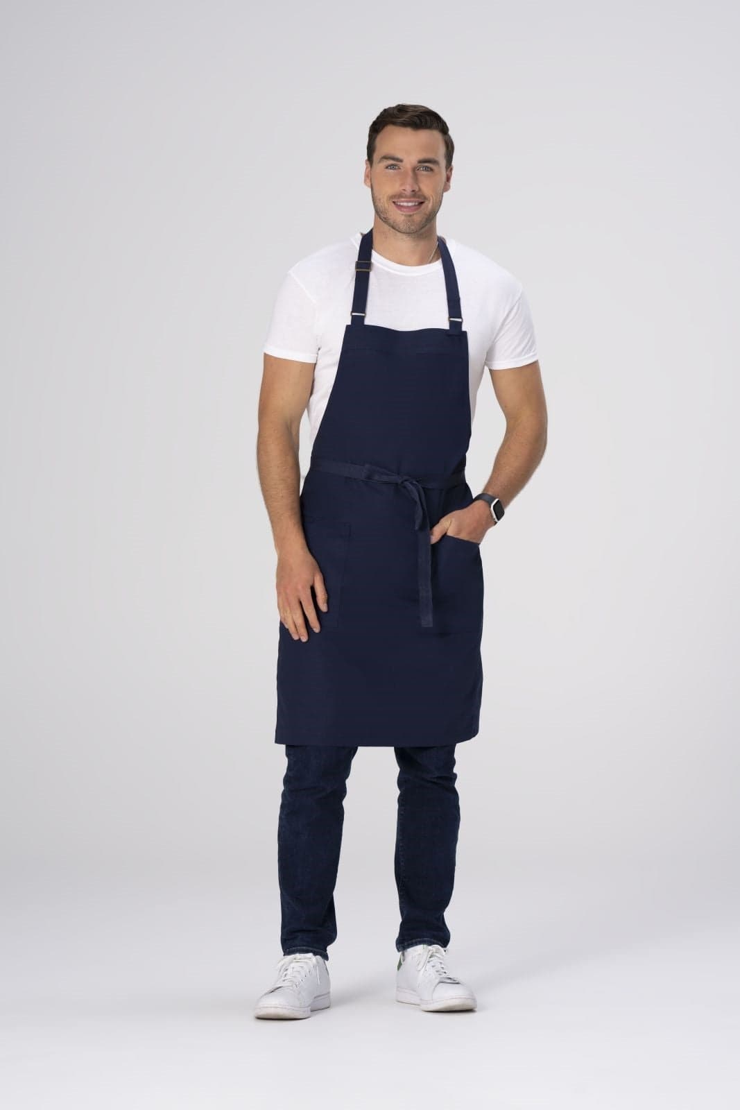 A navy blue bib apron with adjustable straps, worn by a smiling man in casual clothing and white shoes.