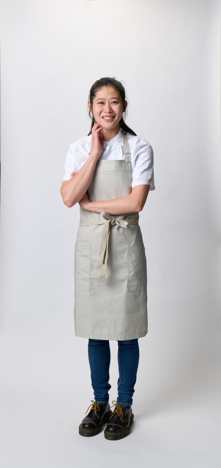 A woman wearing a light grey Lockharte bib apron, paired with a white shirt and dark blue jeans, smiles confidently.