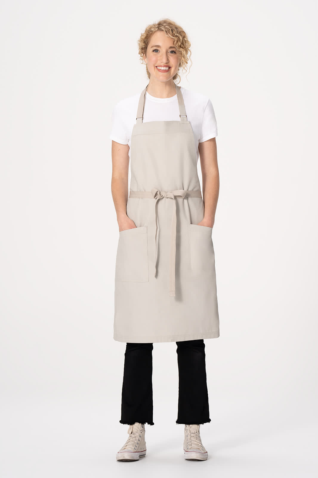 A light beige bib apron with a tie waist and front pockets, worn by a model against a plain background.