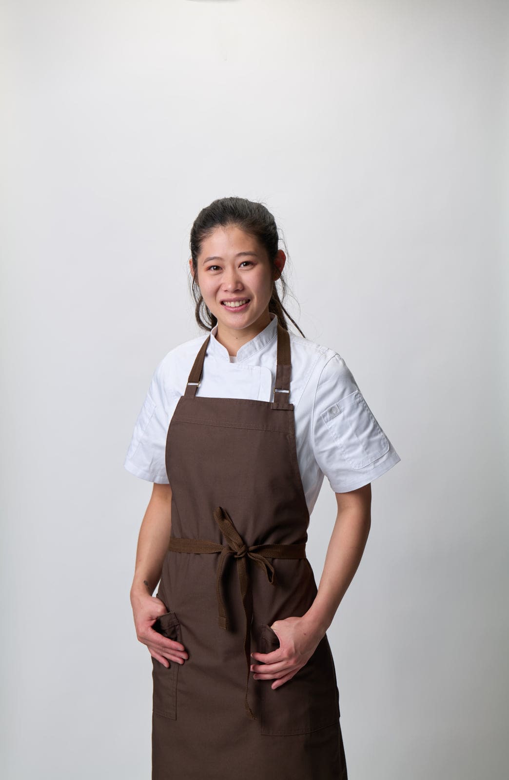 A person wearing a brown bib apron over a white shirt, standing against a light background. The apron has a logo.