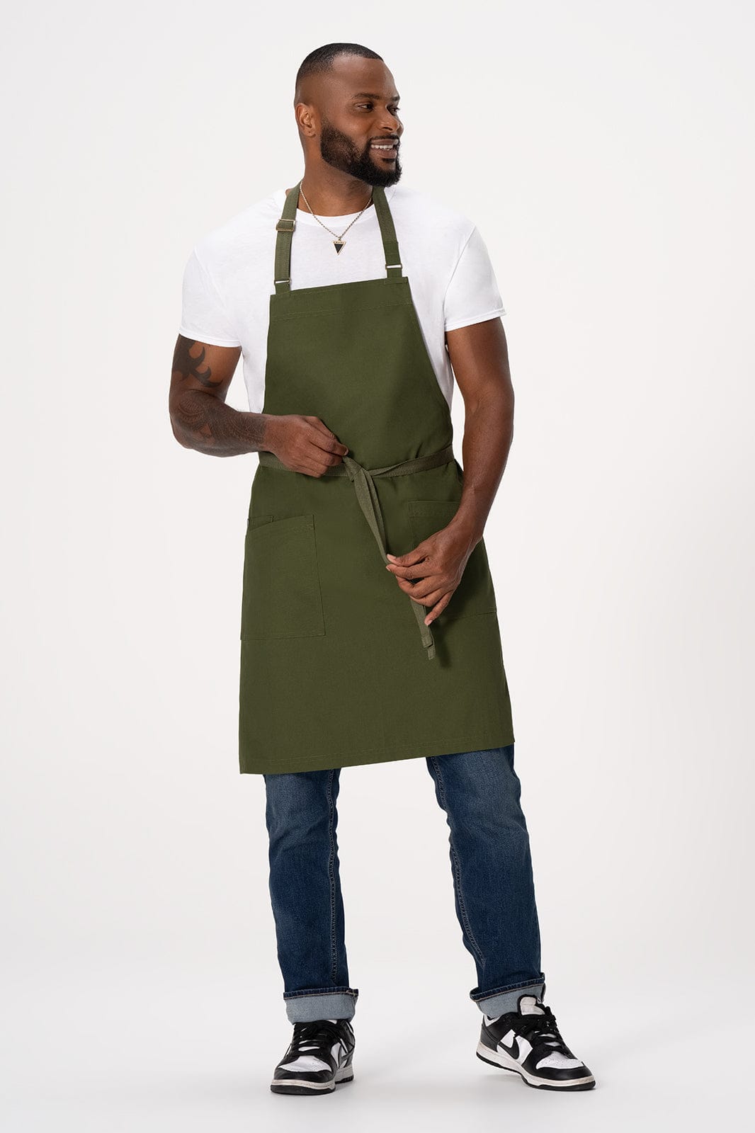 An olive green bib apron is worn by a man, paired with a white t-shirt and blue jeans. The apron has adjustable straps and a logo.