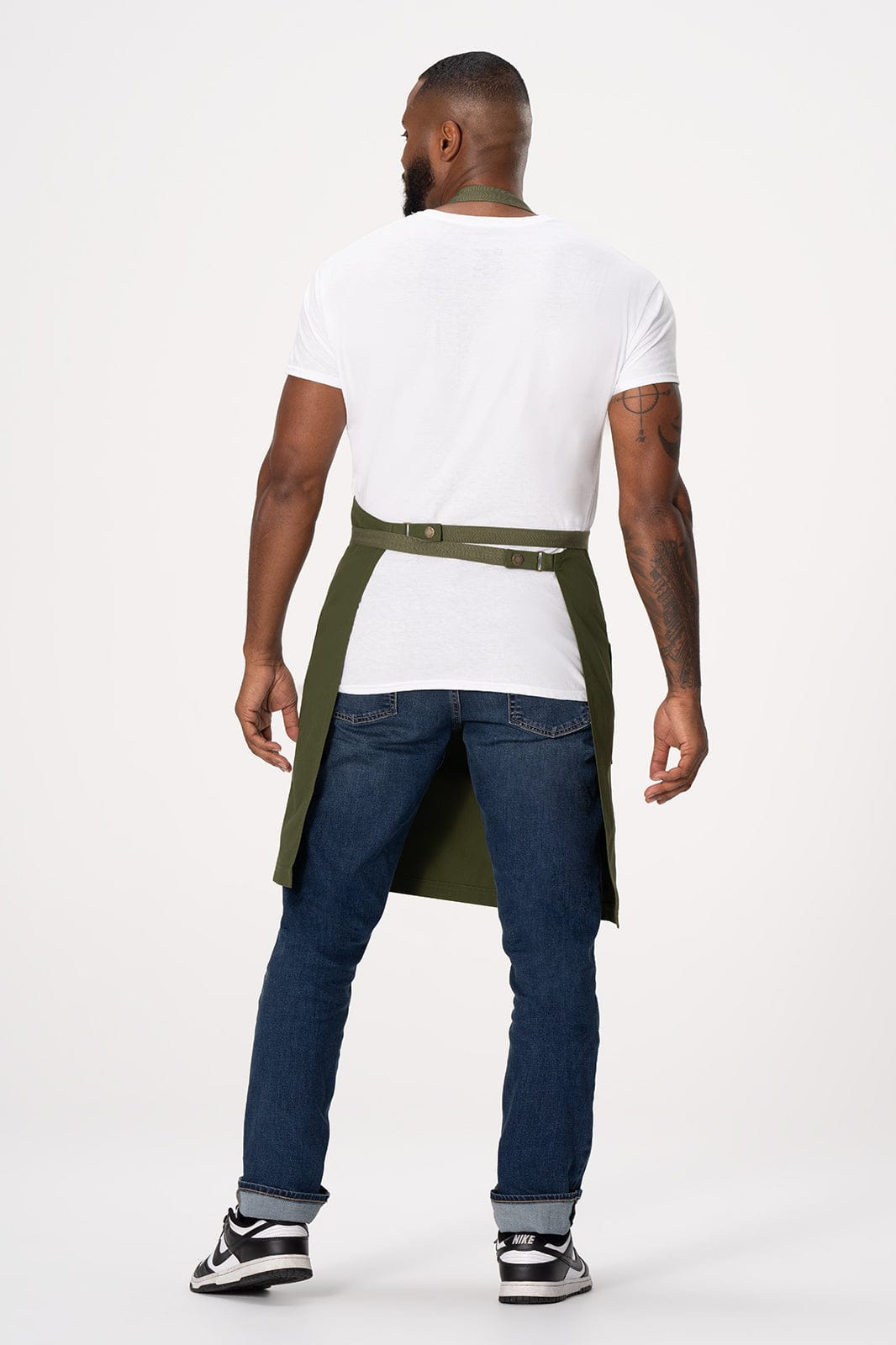 A man wearing a Lockharte Bib Apron in olive green, paired with a white t-shirt and blue jeans. The apron features a logo.