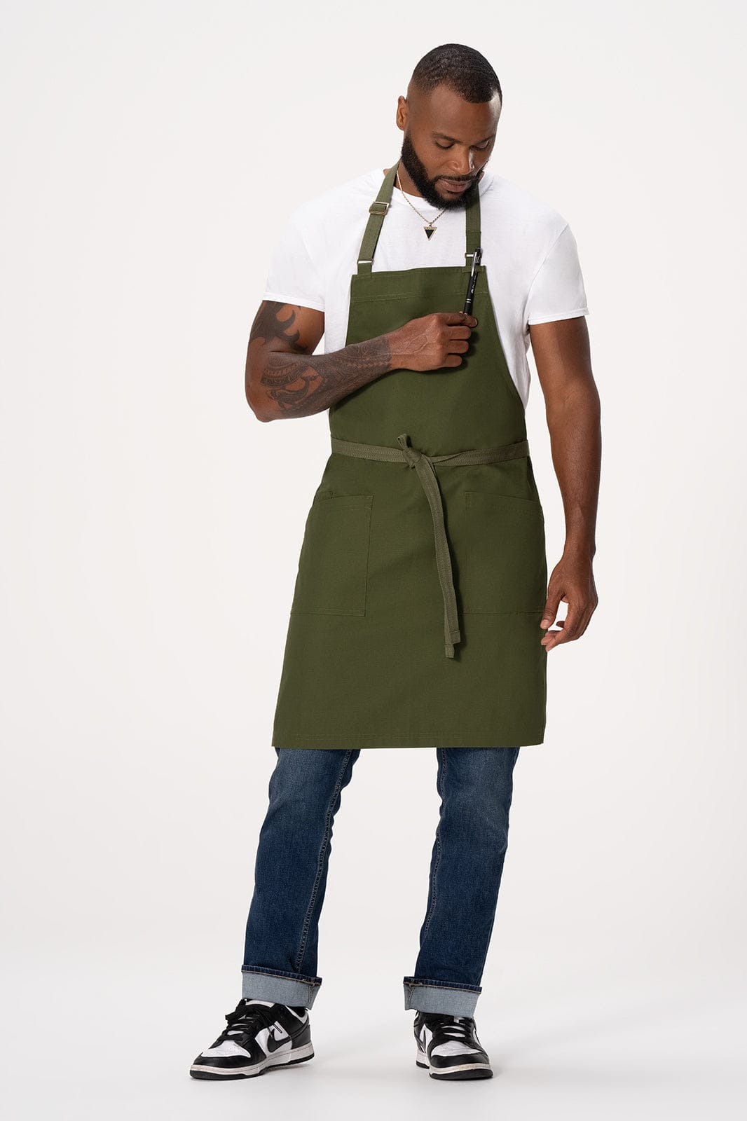 A man wearing an olive green Lockharte Bib Apron over a white T-shirt and jeans, displaying a front pocket.