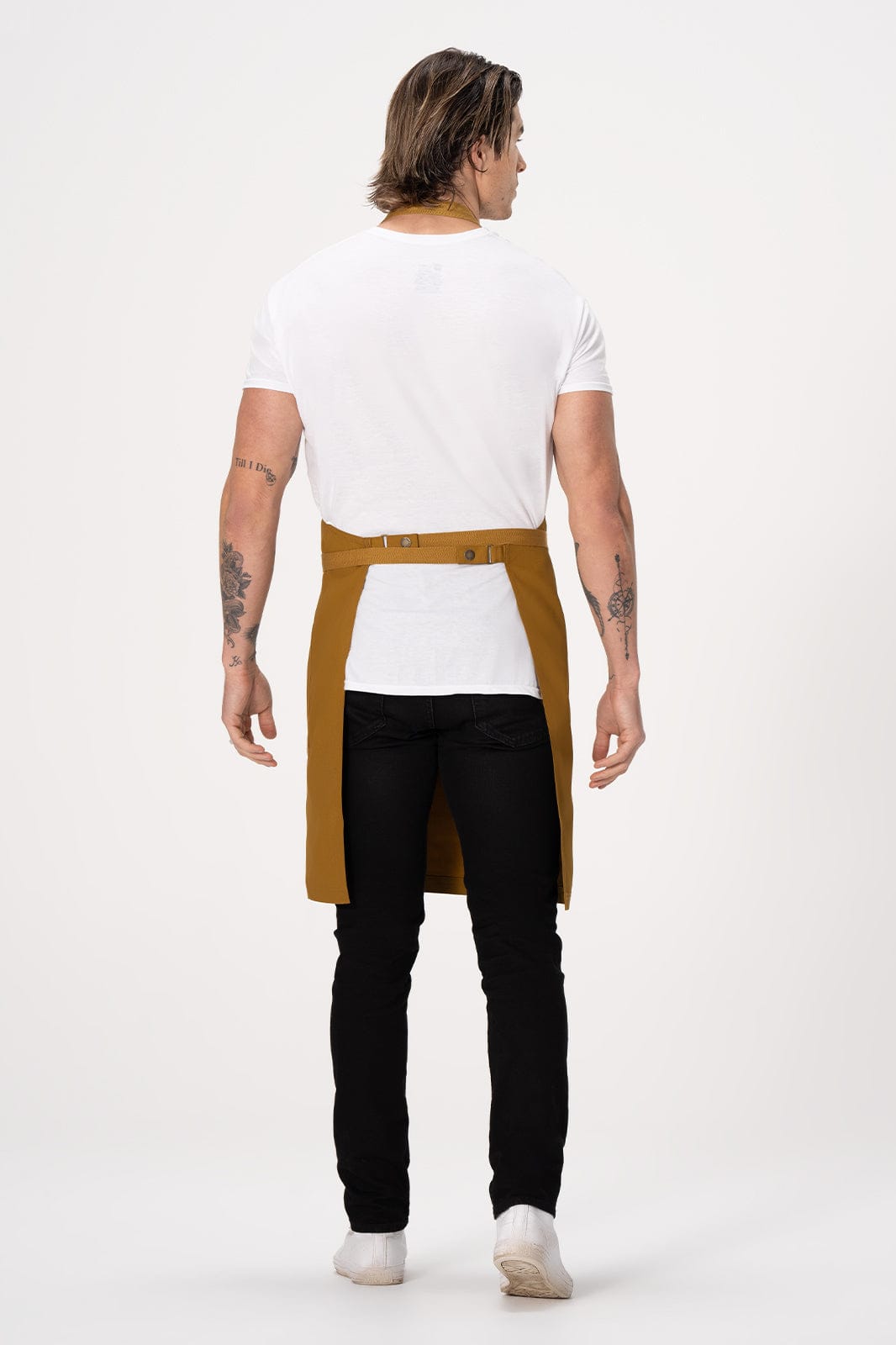 A man standing with his back to the camera, wearing a mustard-coloured Lockharte bib apron over a white shirt and black jeans.