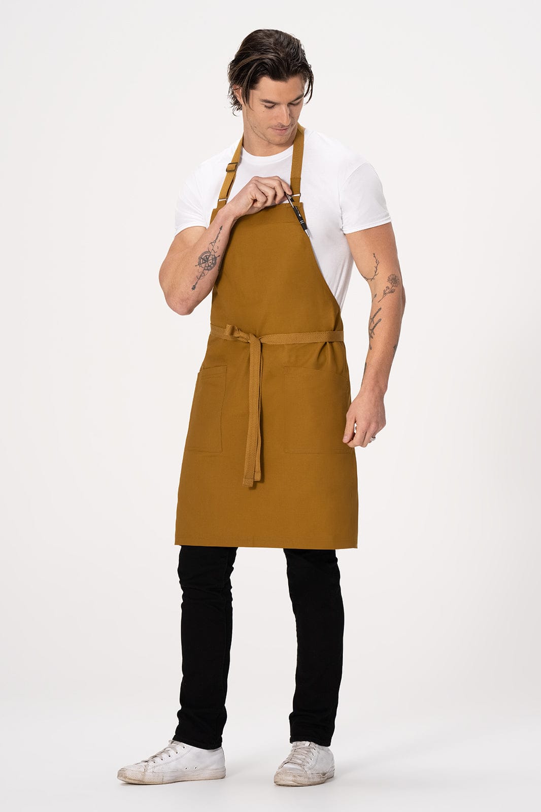 A male model wears a mustard yellow Lockharte Bib Apron over a white t-shirt and black pants, featuring a logo.
