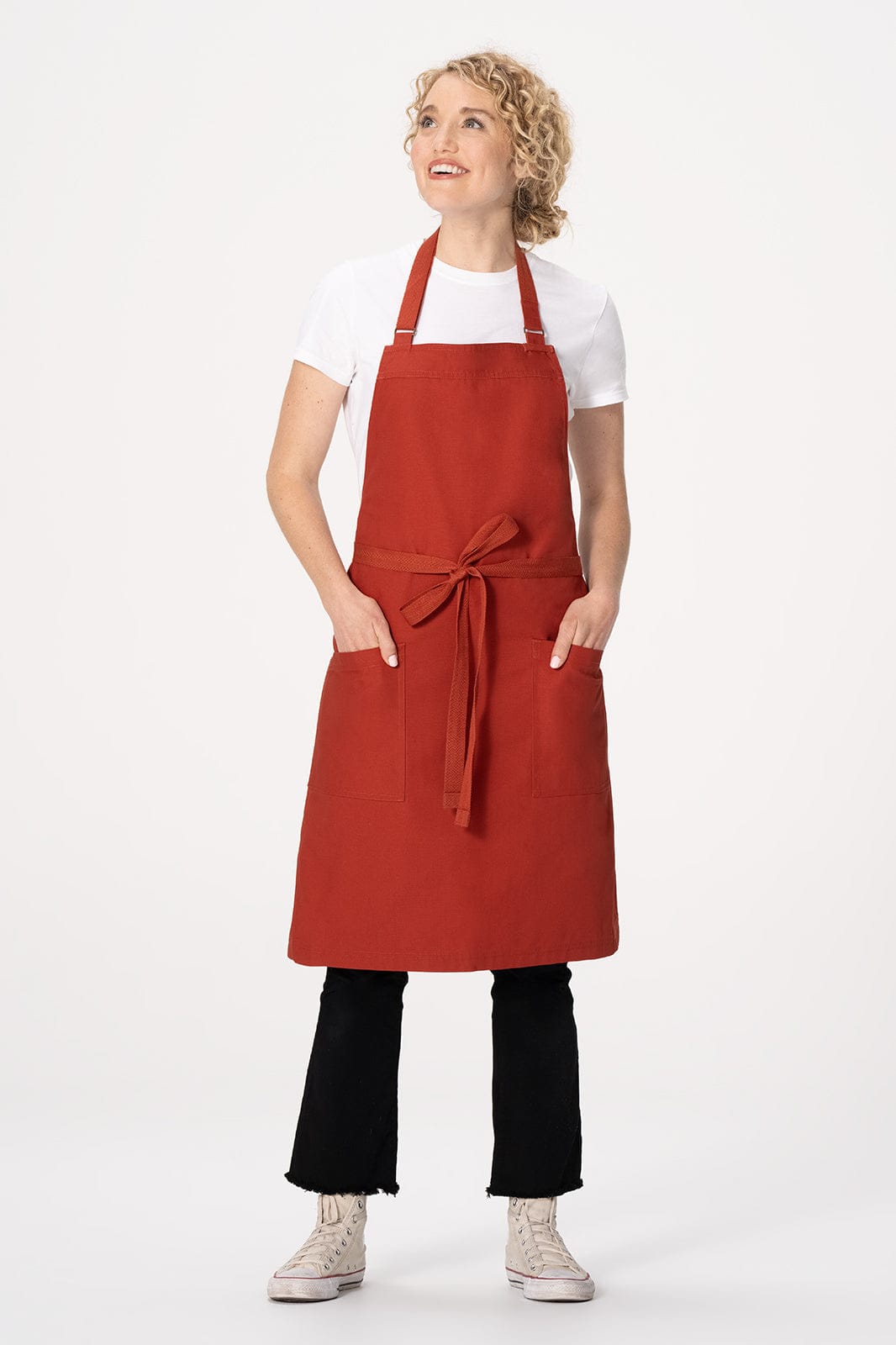 A woman wearing a red Lockharte bib apron, featuring pockets, styled with a white t-shirt and black pants.