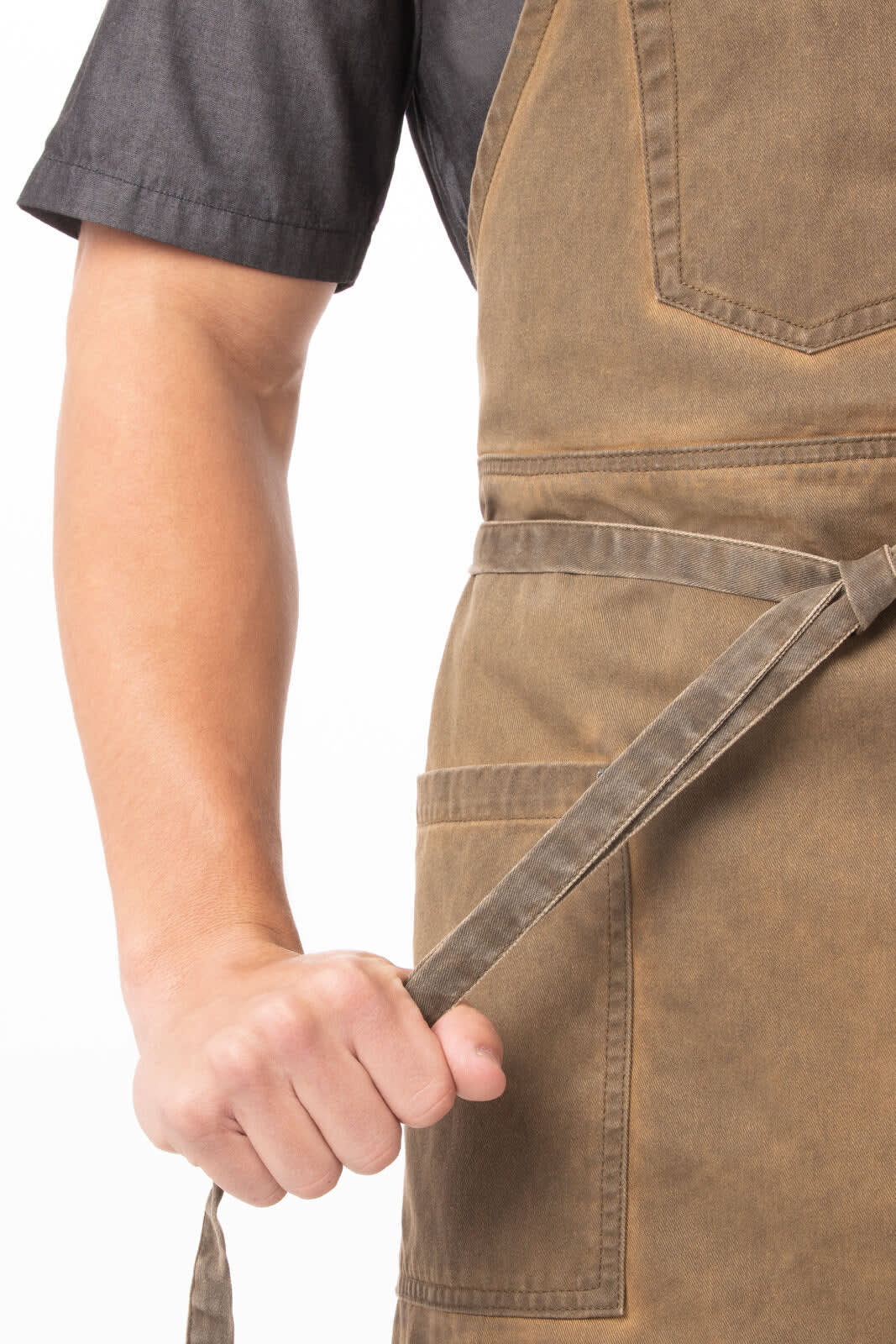 A person in a dark shirt holds the tie of a brown Dorset Bib Apron featuring two front pockets and a logo.