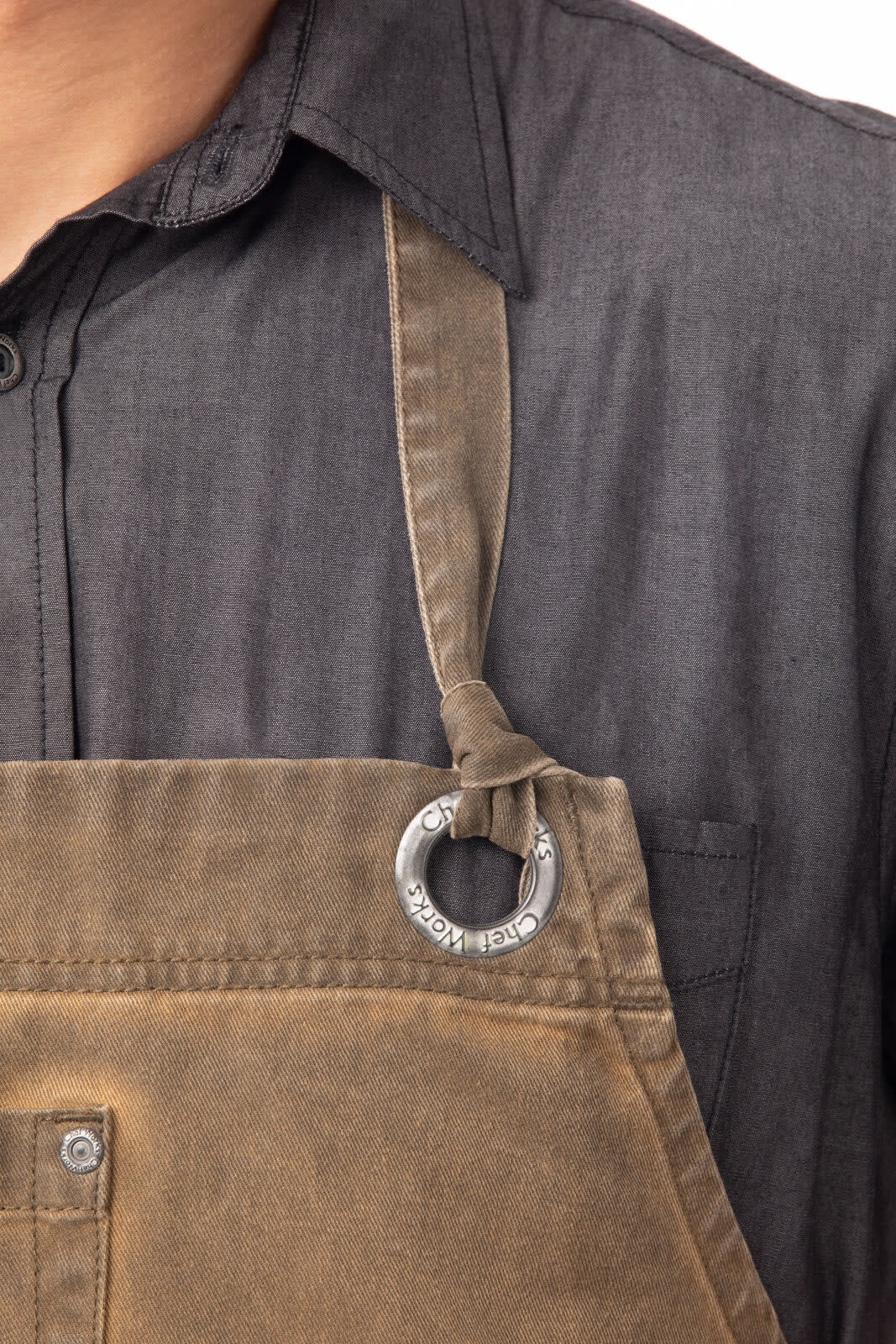 A close-up of a brown Dorset bib apron featuring a metal ring attachment, worn over a black shirt.