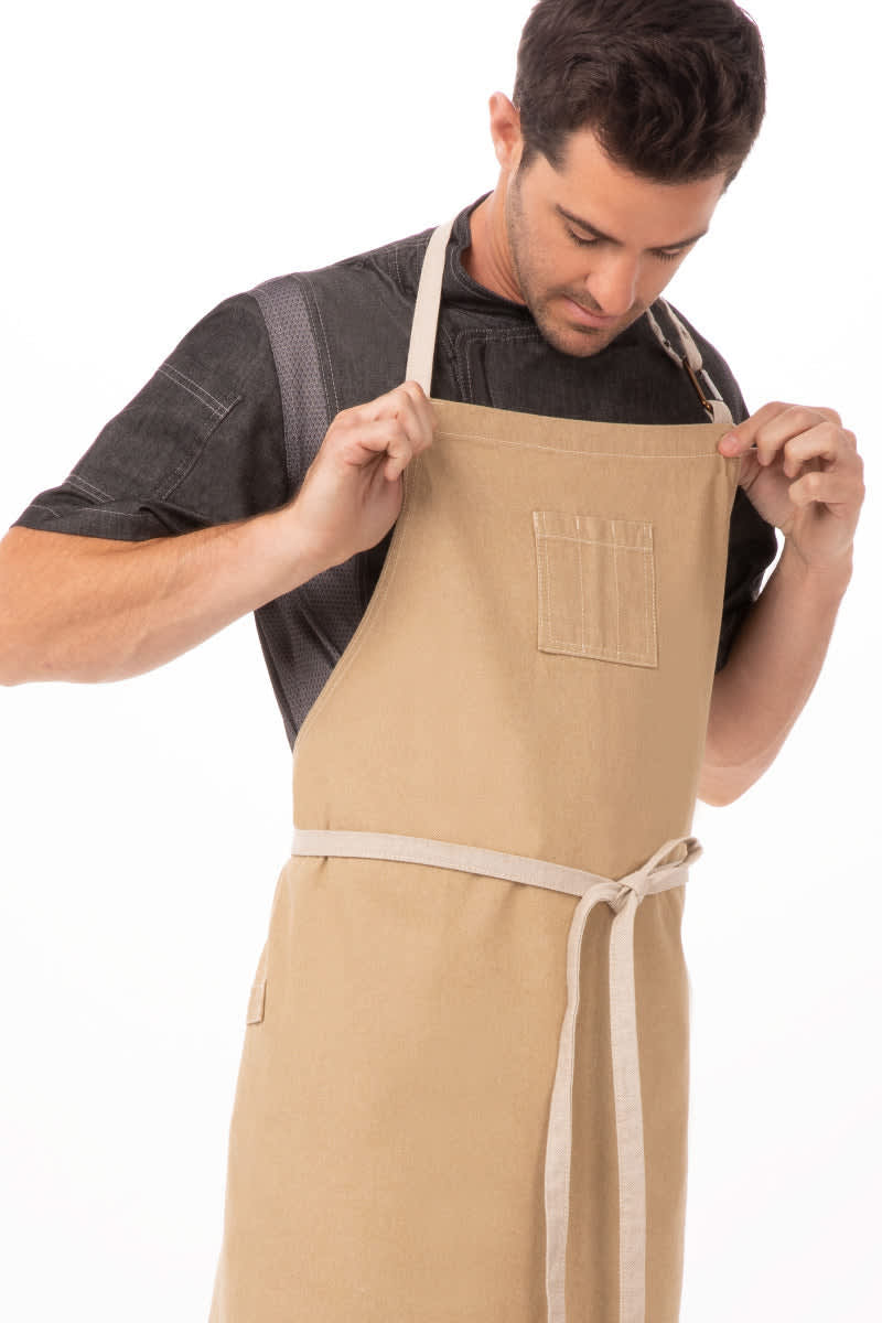 A tan bib apron with a waistband and a chest pocket, worn by a man adjusting it. The apron features branding.