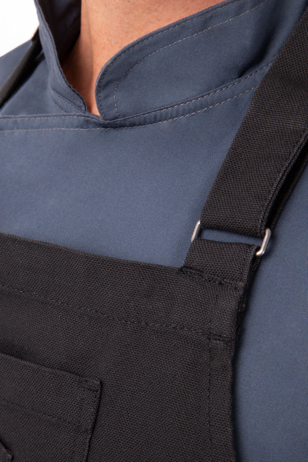 A black bib apron with adjustable straps, worn over a blue chef's jacket. Features a stitched pocket in the front.
