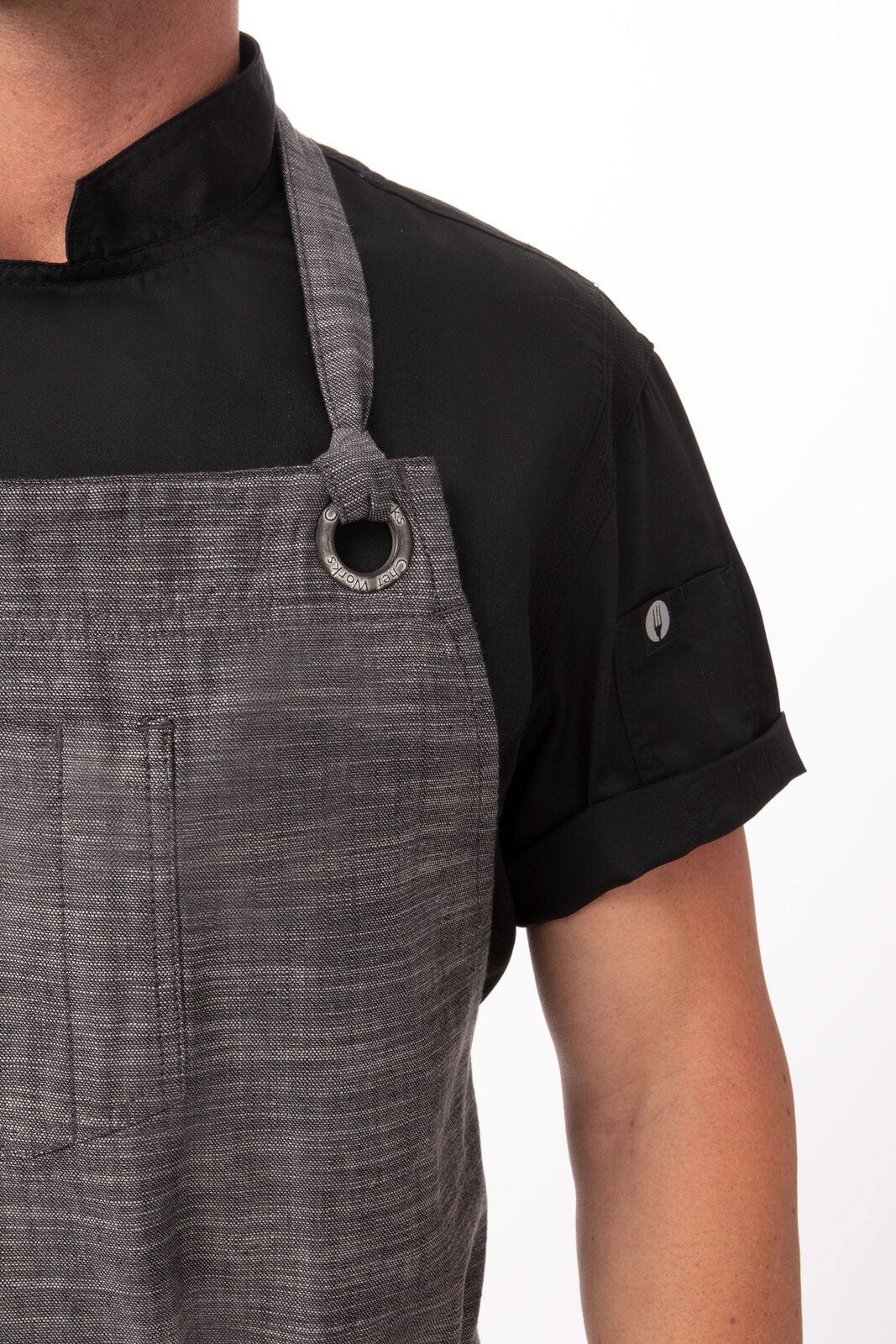 A grey chef's bib apron featuring a grommet detail, worn over a black shirt. Includes a pocket and logo branding.