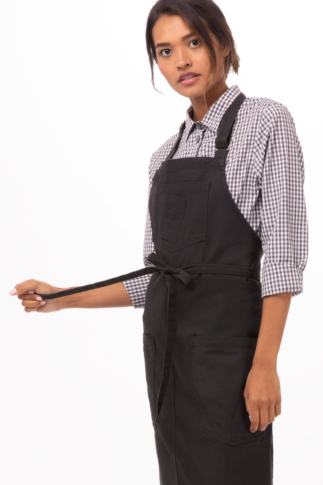 A woman in a black bib apron over a checked shirt, holding the apron ties. The apron features a pocket and a logo.