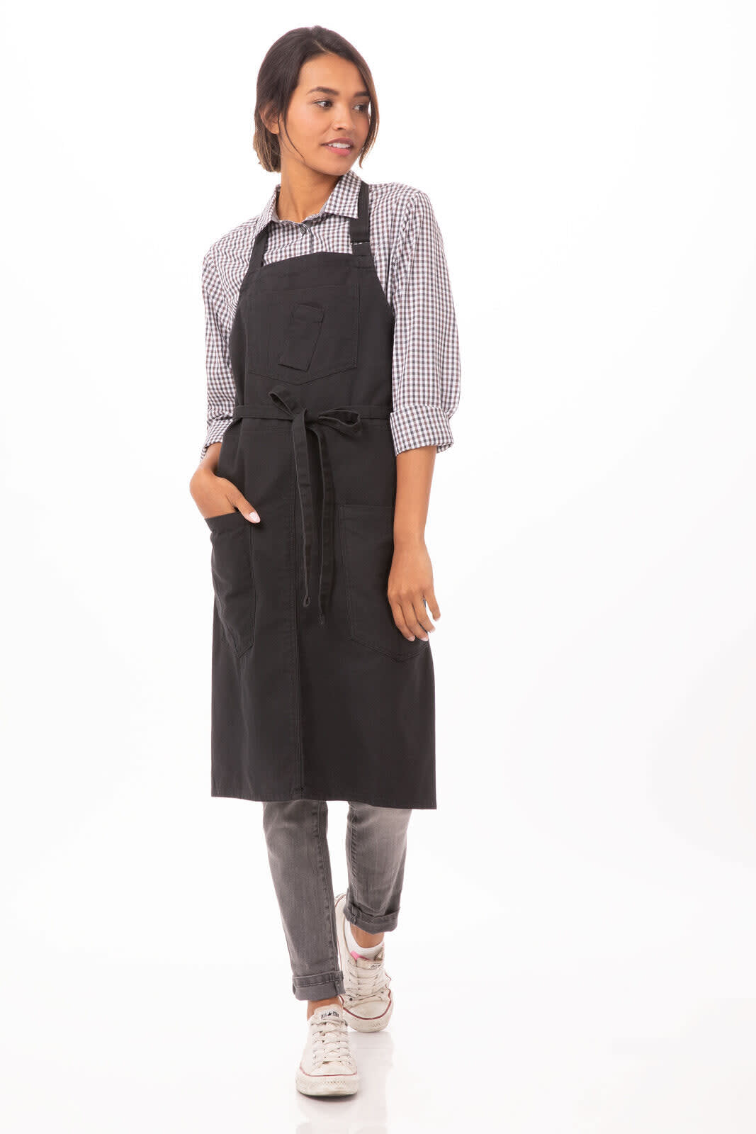 A black bib apron is worn by a person with a checkered shirt and grey jeans, featuring a pocket and a tied waistband.