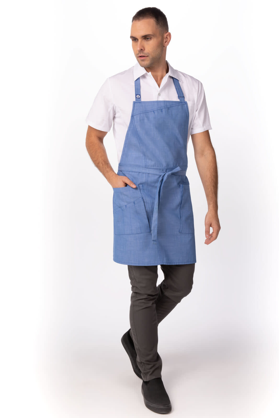 A blue bib apron worn by a model, featuring pockets and an adjustable neck strap, paired with a white shirt and dark trousers.