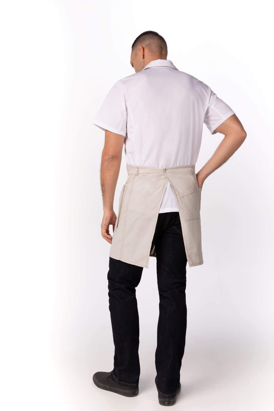 A man wearing a beige Medford Bib Apron, standing with his back towards the viewer, paired with dark jeans.